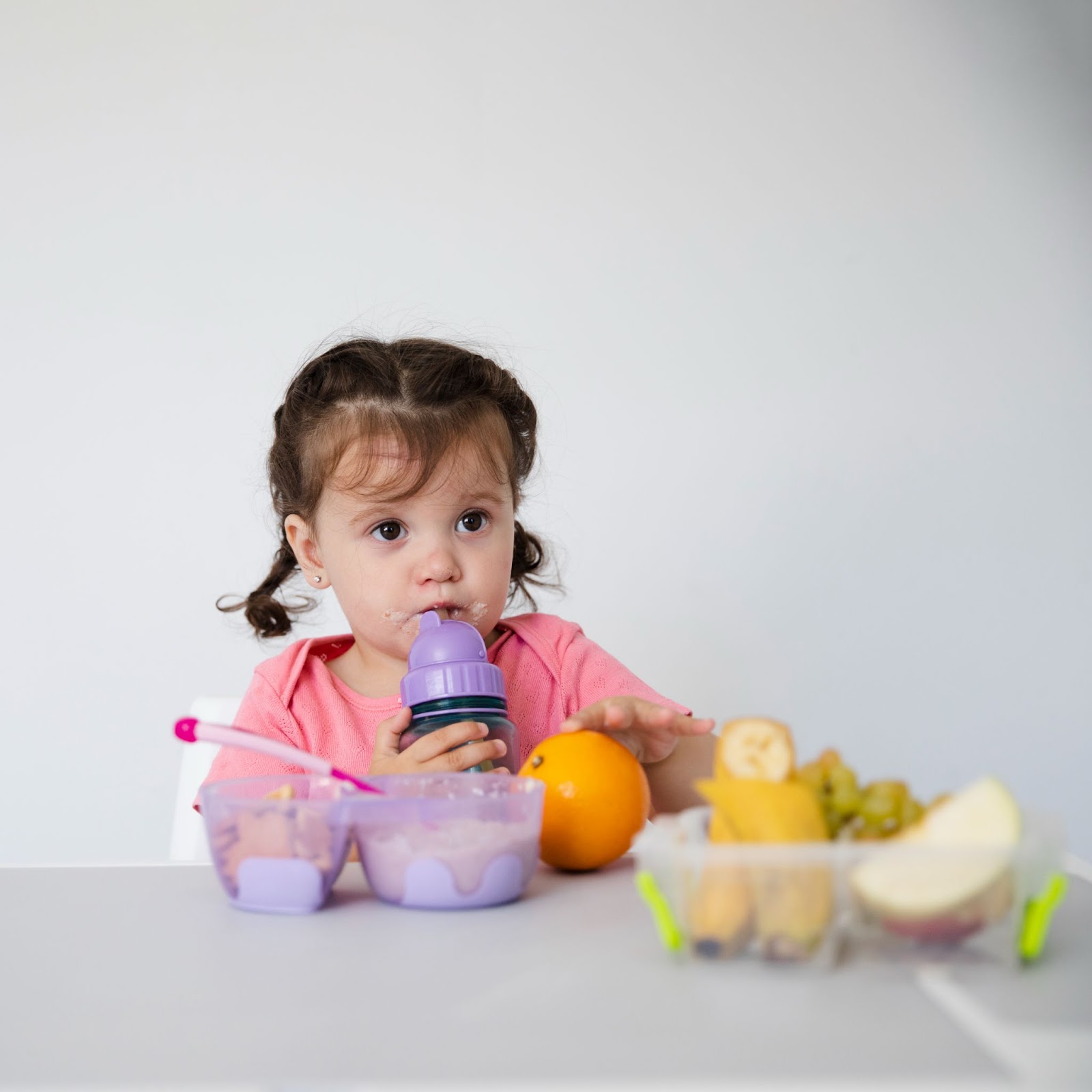 A little girl sipping her water with a fruit bowl in front