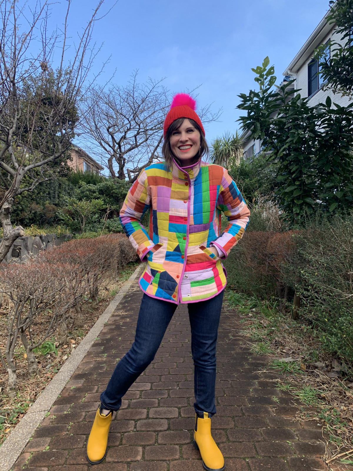 a white woman stands on a path outside wearing a quilted jacket composed of brightly colored fabric scraps