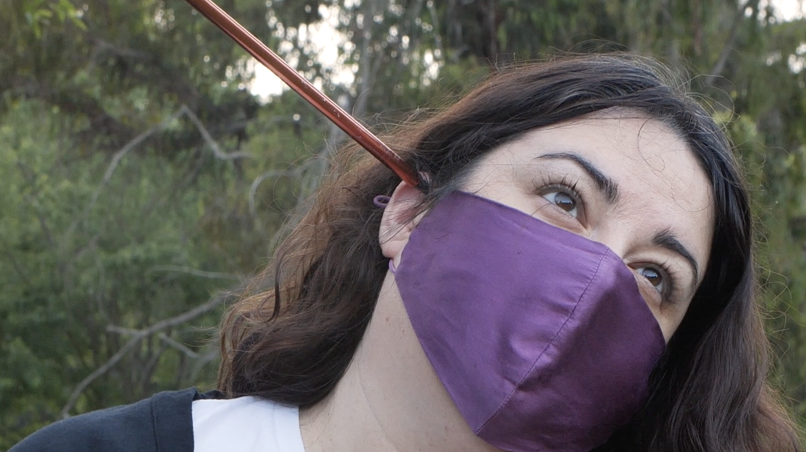 a photo of Selwa, an arab american woman with light skin, dark eyes, round face, and shoulder length wavy dark and gray hair. She is looking upwards, and is wearing a satiny purple face mask, while leaning to the right of the frame. A metallic prop is resting on her ear. She is outdoors, it is daytime and in the background are trees and green brush.