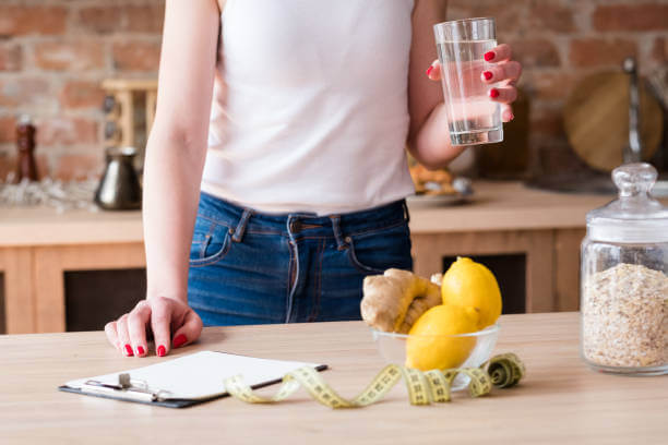 A person standing at a kitchen counter preparing a detox drink with fresh ginger, lemons, and a glass of water.