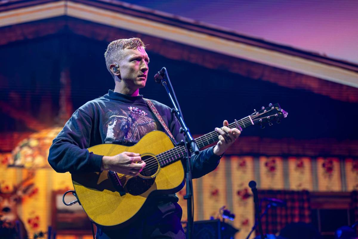 A white man holds an acoustic guitar on stage with a barn-home backdrop