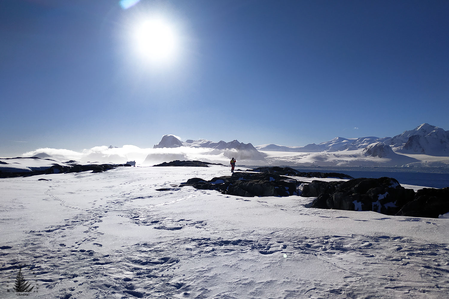 An image of a single human walking along a sunlit, snowy surface with a bright blue sky in the background.