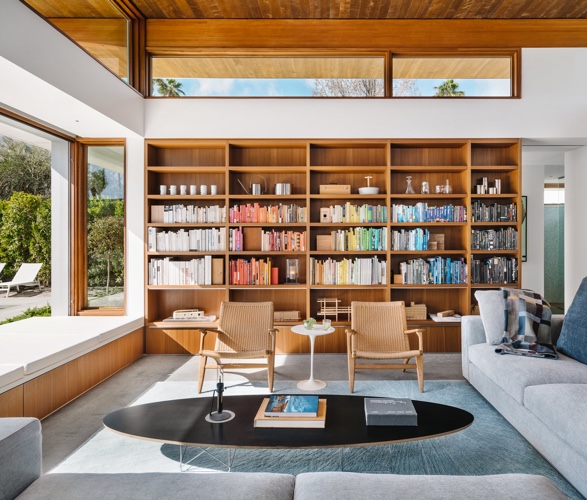 A mid-century modern living room with floor-to-ceiling wood bookshelf organized by color, large windows and minimalist lounge chairs.