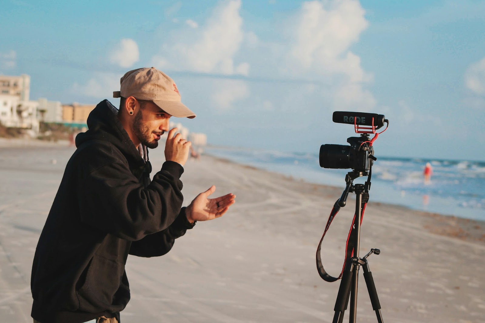 A man infront of a camera on a sunny beach with waves in the background.