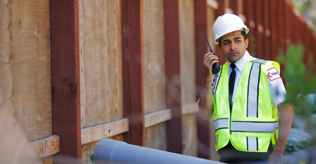 (A security guard in a high-visibility vest and hard hat communicates via radio while patrolling a construction site perimeter, ensuring safety and protection.)