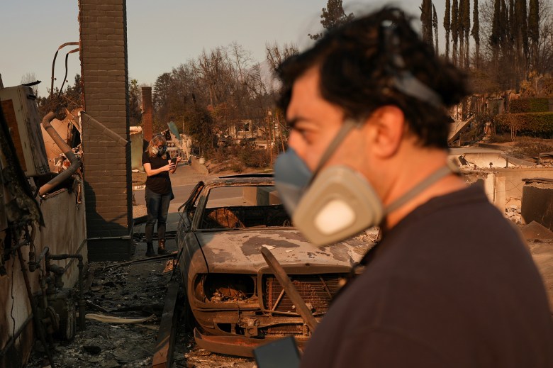 Foreground shows an individual wearing a respirator mask looking downward. In the background, another person in a mask examines the scene using a mobile phone. A burned-out car sits amidst the charred remnants of a structure, with trees and other fire-damaged areas visible in the distance under a hazy sky.