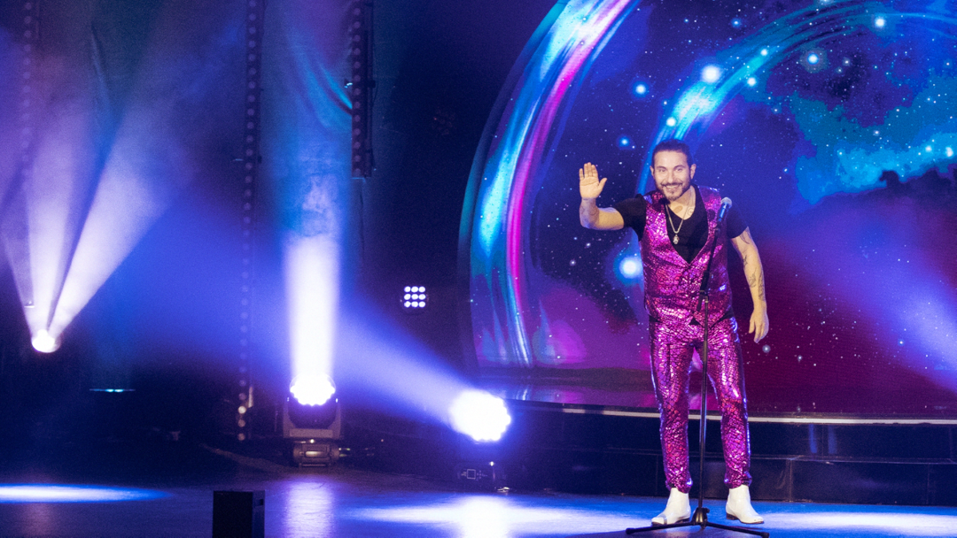 Hombre vestido con traje brillante de color morado saludando en un escenario iluminado con luces de colores y fondo galáctico.