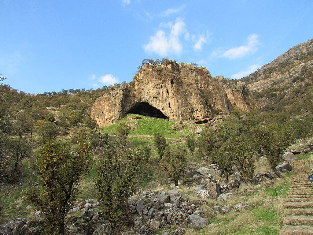 View of Shanidar Cave in the Kurdistan Region of Iraq, showcasing its large rock opening and surrounding natural landscape with trees and rocky terrain.