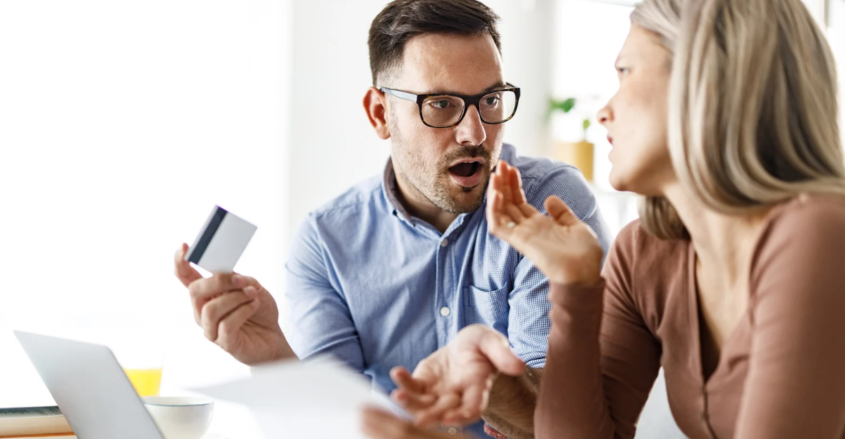 (A couple having an intense discussion at a table, with the man holding a credit card and the woman gesturing, surrounded by papers and a laptop.)