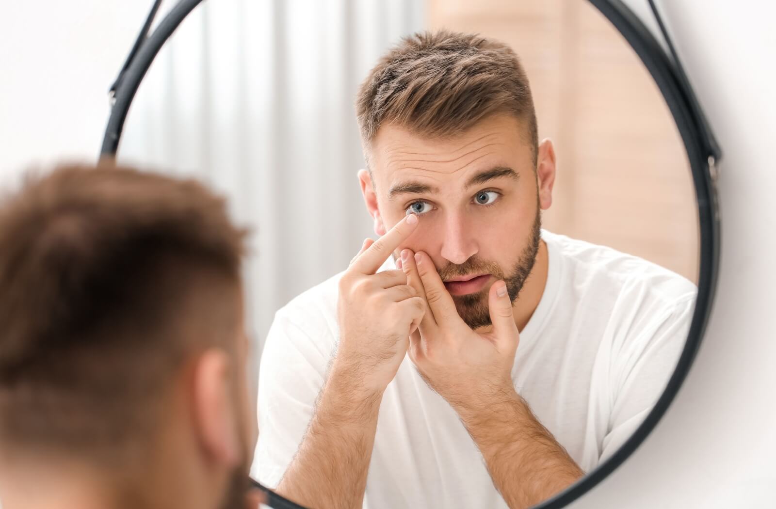 A young man looking in a mirror and putting a contact lens in his left eye.