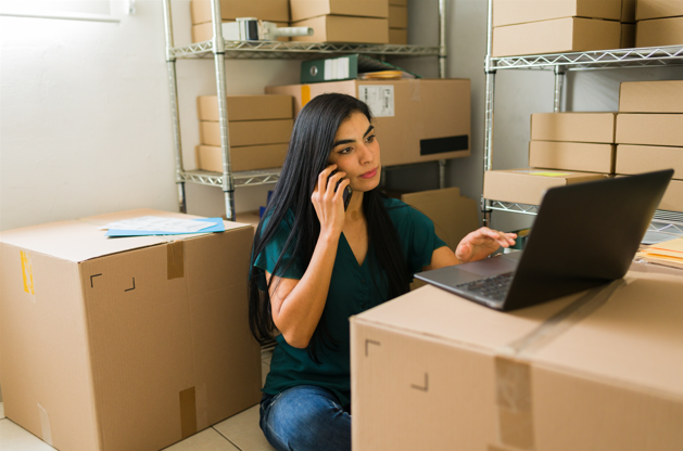 Mujer en almacén, llamando teléfono y consultando su computadora para resolver problemas logísticos B2B. 