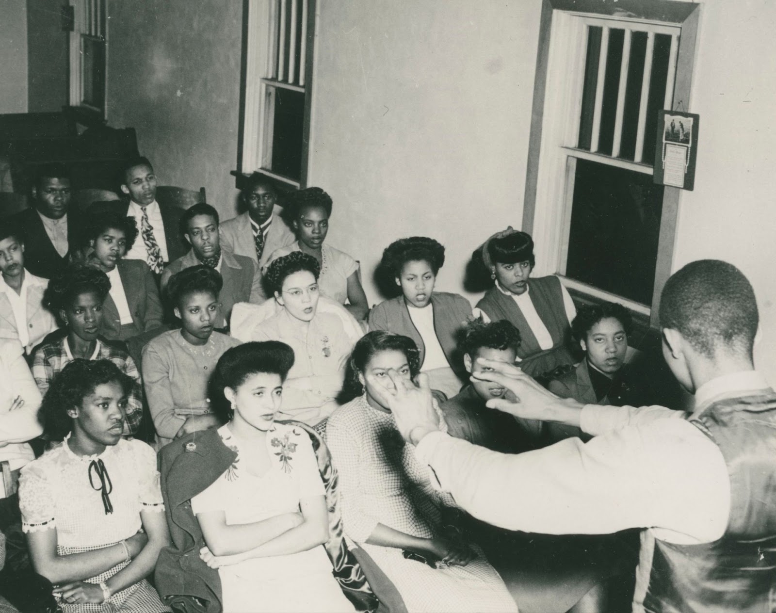 A choir sits in front of a man to the right of the image as he directs them with his hands. 
