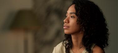 A portrait photograph of a black woman with long permed hair wearing beige clothing, looking relaxed as she gazes towards the left side.