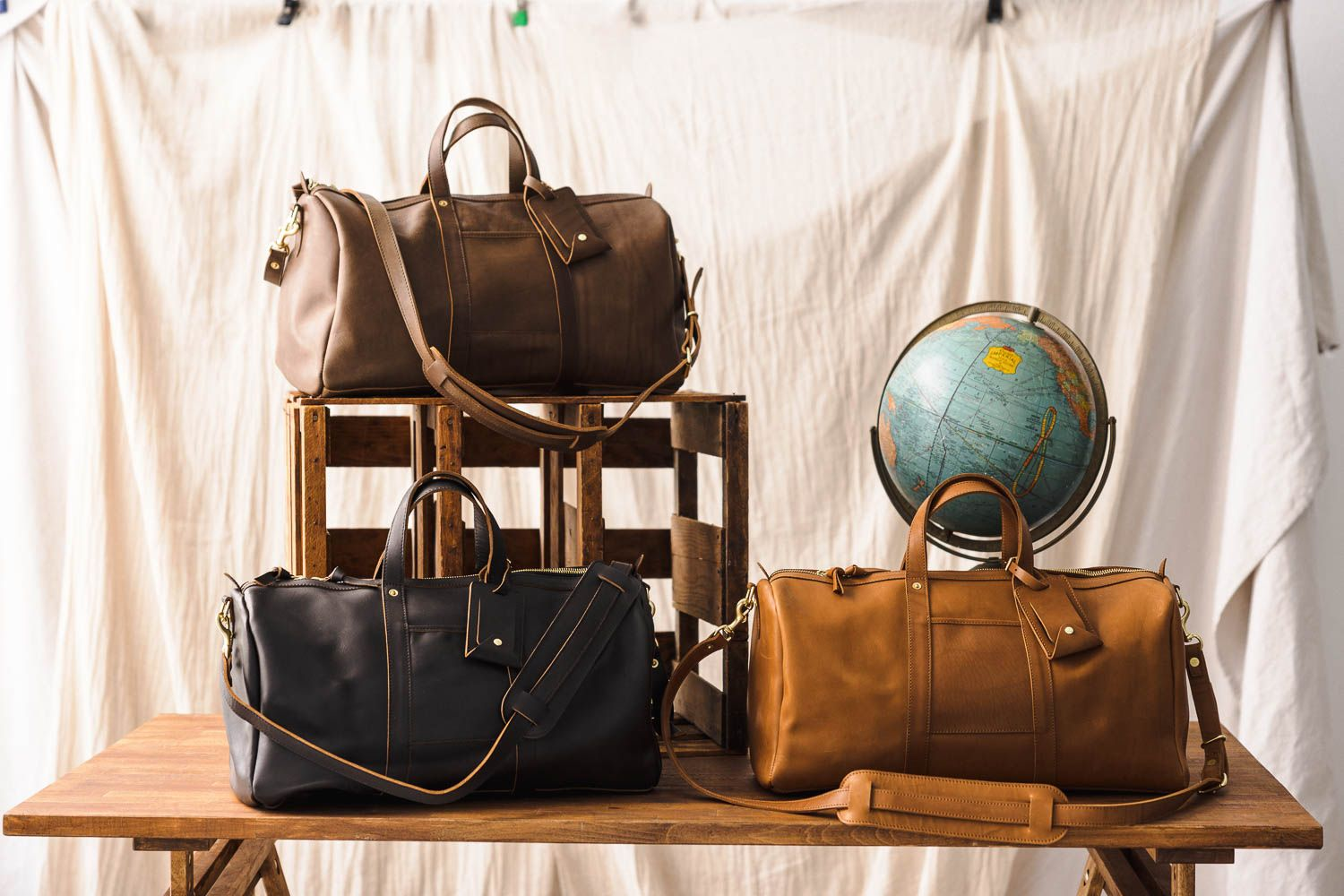 Three leather duffel bags from WP Standard in different colors are displayed on a wooden table, with a globe in the background.