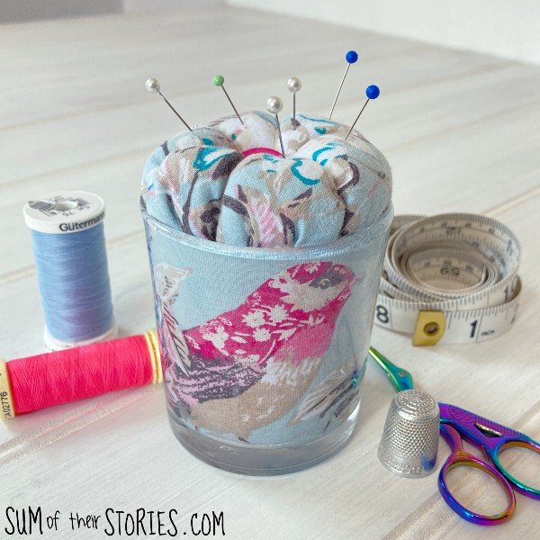 sewing notions on a white wood table next to an upcycled candle jar covered in fabric and topped with a fabric pin cushion cover
