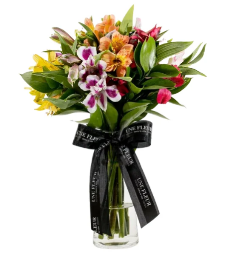 Alstroemérias coloridas com folhagens verdes em um vaso de vidro alto transparente com água até a metade e um laço preto com a logo da Une Fleur entrelaçando o vaso.