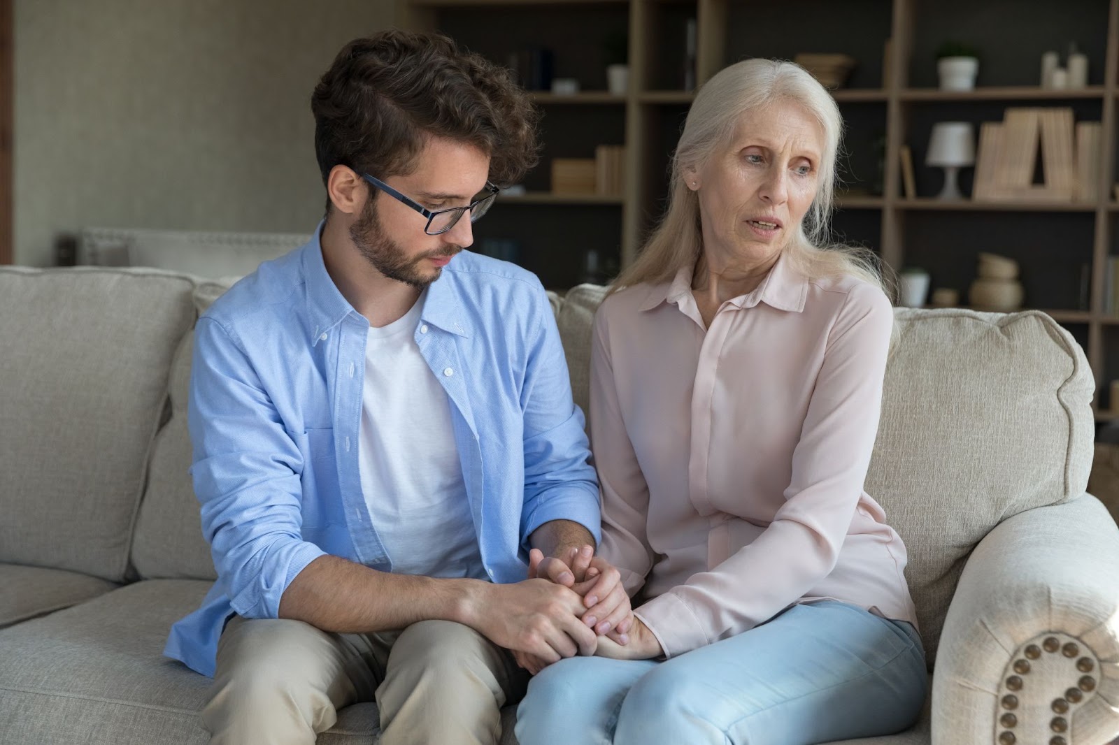 An adult son sitting next to his mom on the couch holding her hands to comfort her as she looks away.
