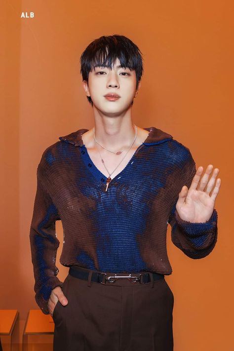 This contains a picture of BST's JIN  standing in front of an orange wall holding his hands out to the side