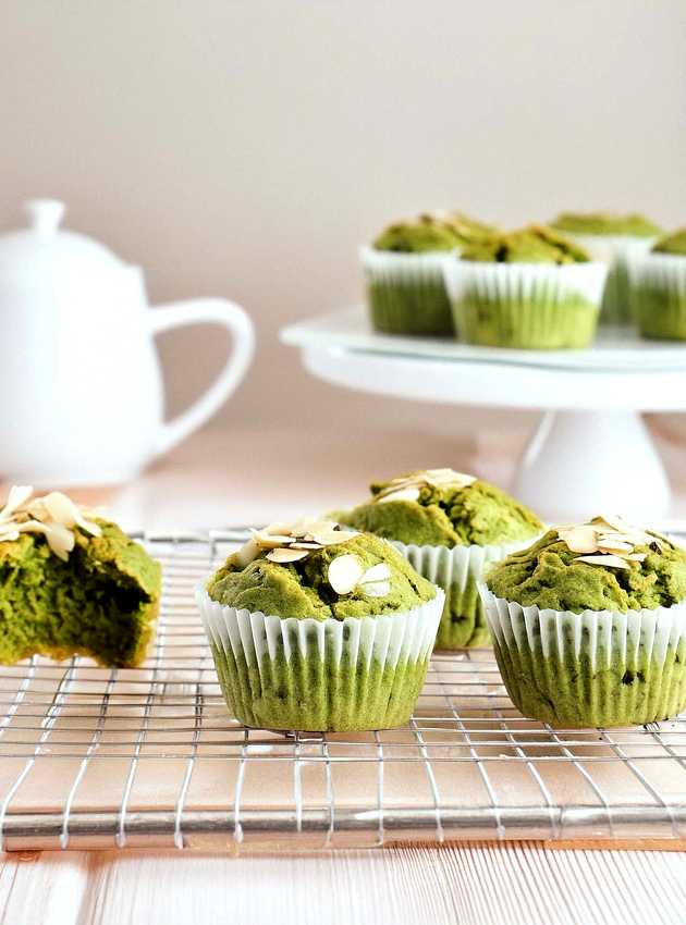 Green muffins with almond slivers cooling on a wire rack, with more muffins on a cake stand in the background.