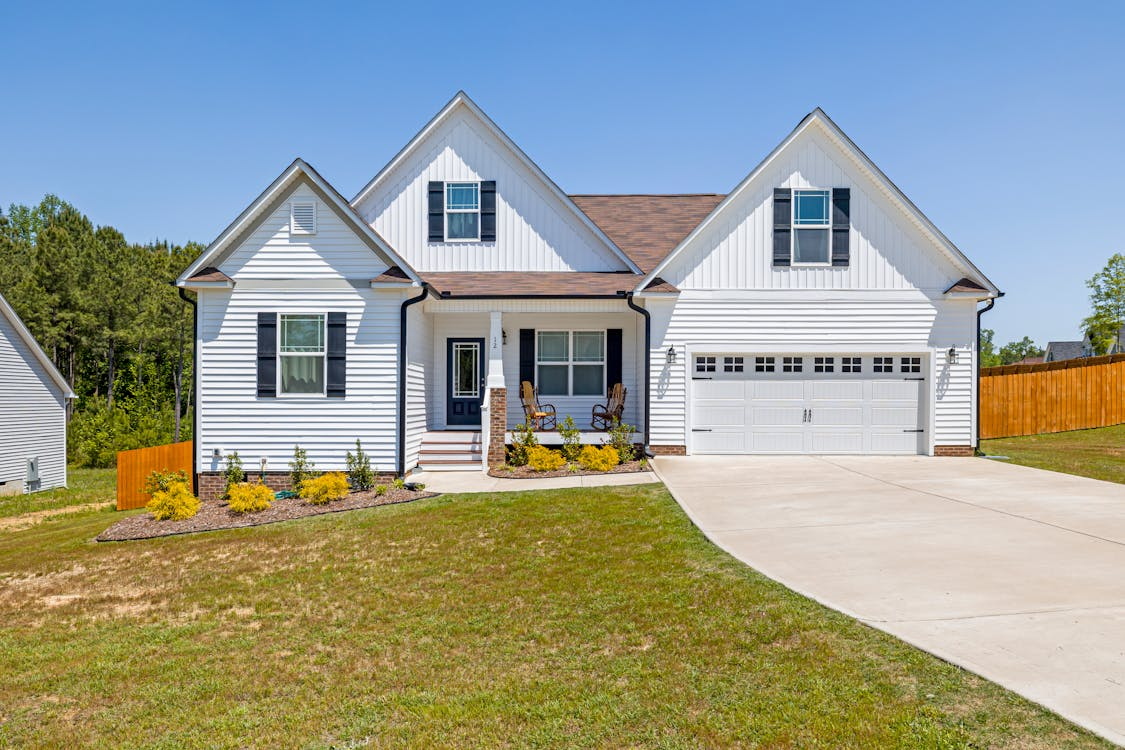 A white single-story house with gable roofs, black shutters, and a large driveway leading to a two-car garage, surrounded by a well-maintained lawn and bordered by a wooden privacy fence under a clear blue sky