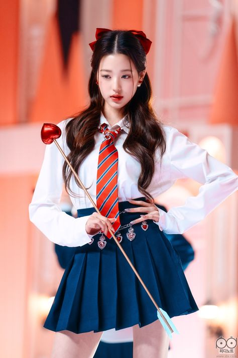 This contains an image of Wonyoung's  in a skirt and tie holding a wand with her hands on her hips