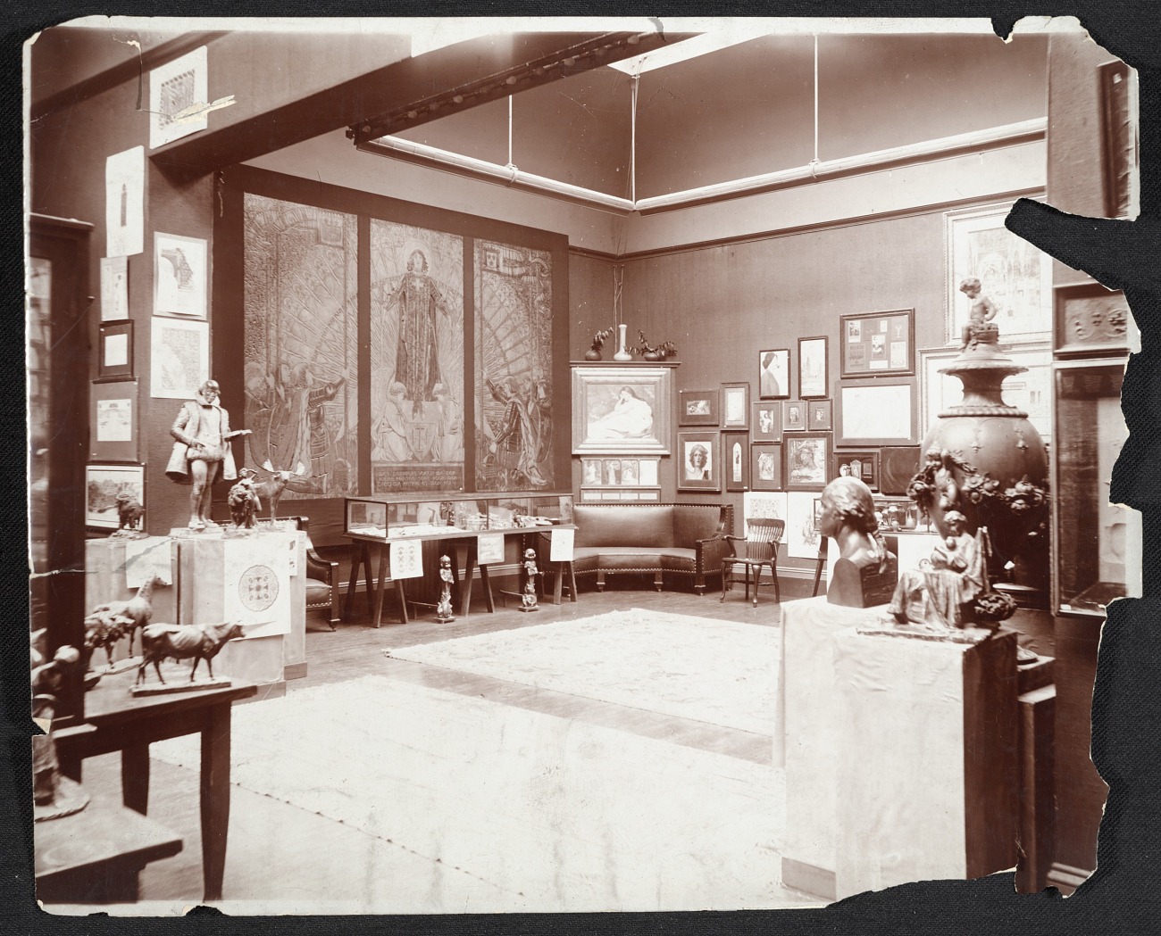 Vintage photograph of a 19th-century art gallery room displaying various sculptures, paintings, and framed artworks on the walls, with ornate vases, busts, and animal figurines arranged on pedestals and tables, and a large mural as the central focus, evoking a classical museum exhibition setting.