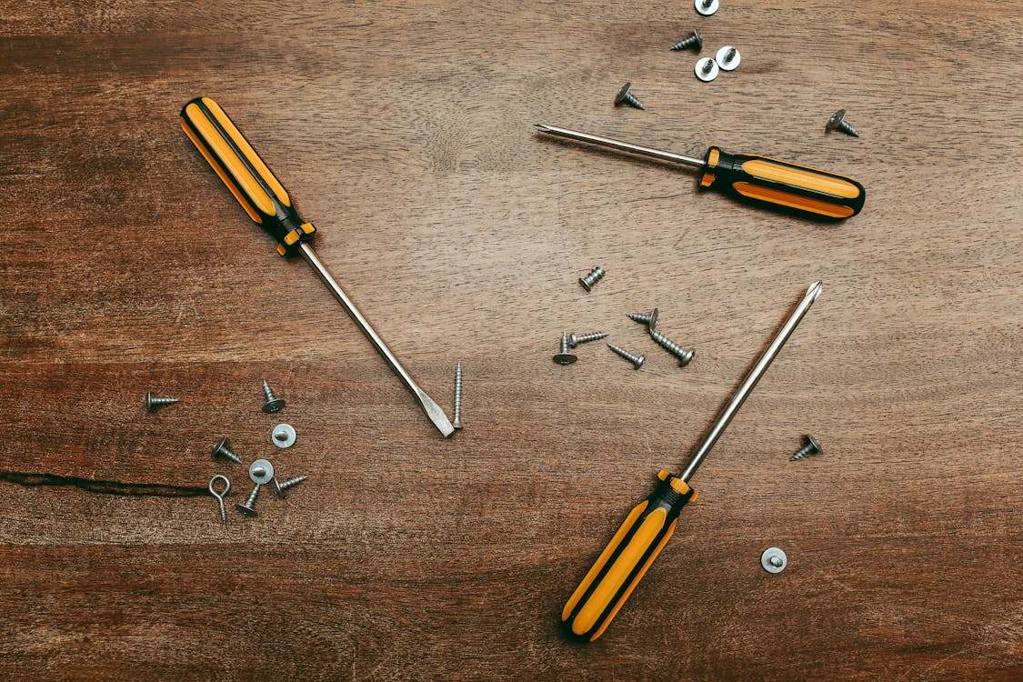 Three screwdrivers with black and yellow handles placed on a wooden surface surrounded by scattered screws and washers, illustrating tools and hardware for assembly or repair