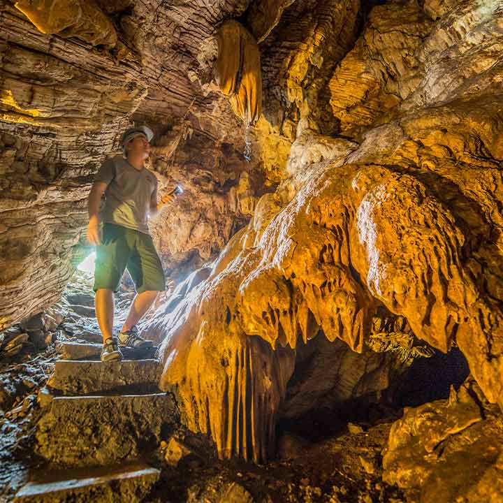 Homem explorador explorando uma caverna de estalactites e estalagites douradas com iluminação natural, vestindo roupas de aventura e usando lanterna.