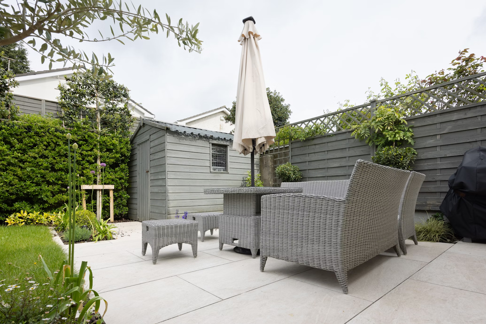 Modern backyard patio with gray wicker furniture, a closed umbrella, tiled flooring, and a painted wooden shed surrounded by greenery and privacy fencing.