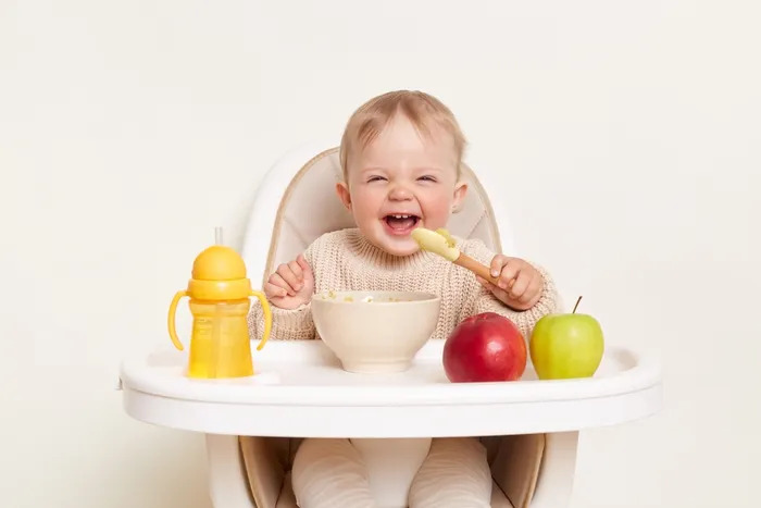 A baby in a high chair, playing with a spoon and surrounded by fresh apples on the tray.