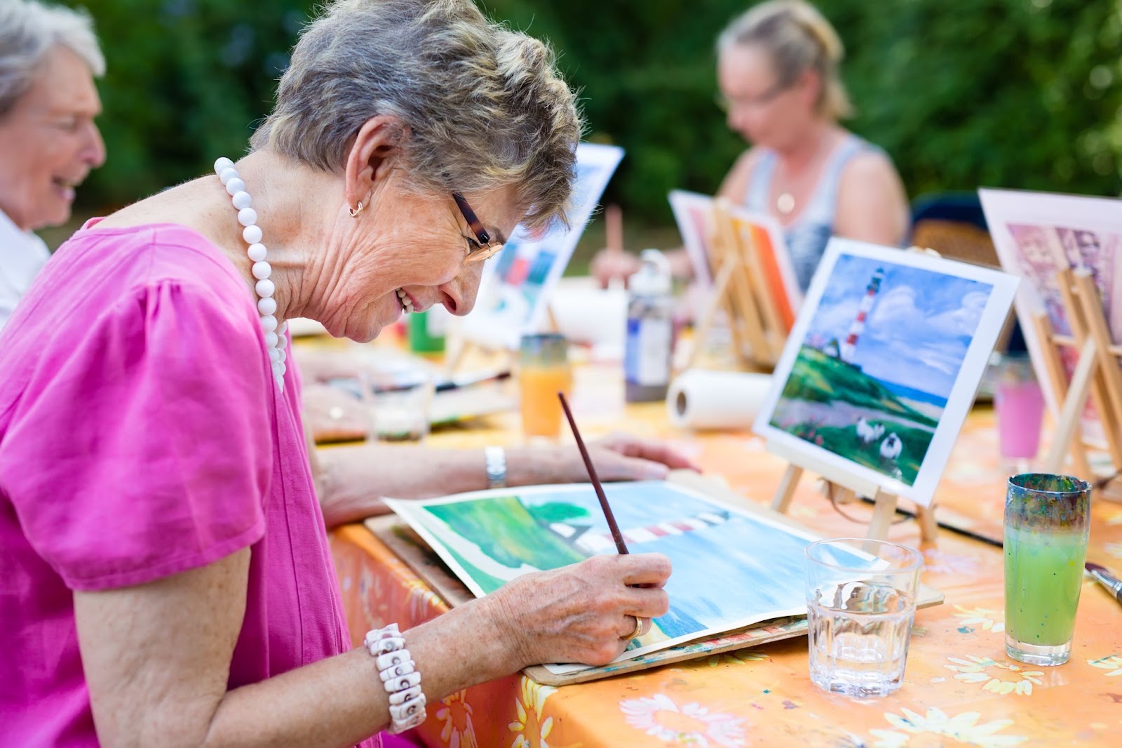 A senior woman in memory care paints a lighthouse on a canvas among her friends.