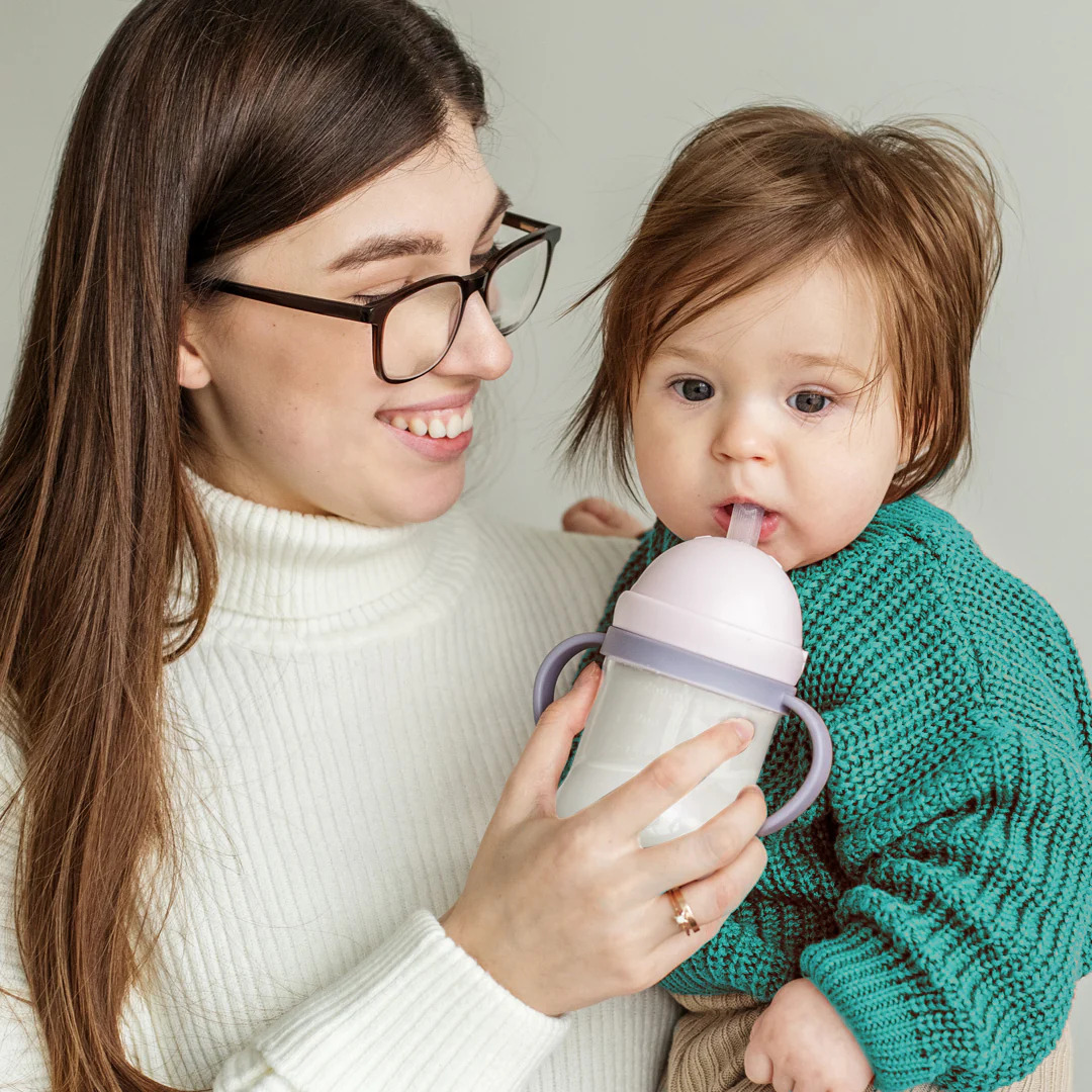 A woman cradles a baby in one arm while drinking from a bottle with the other hand.