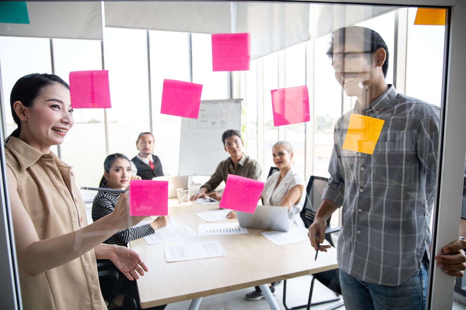 A team of business people meeting in an office, using pink post-it notes on a glass wall to share and discuss ideas for developing a profitable business idea.