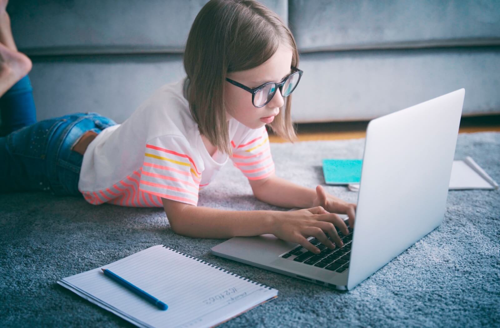 A child wearing eyeglasses uses a computer on their living room floor, school supplies surrounding them