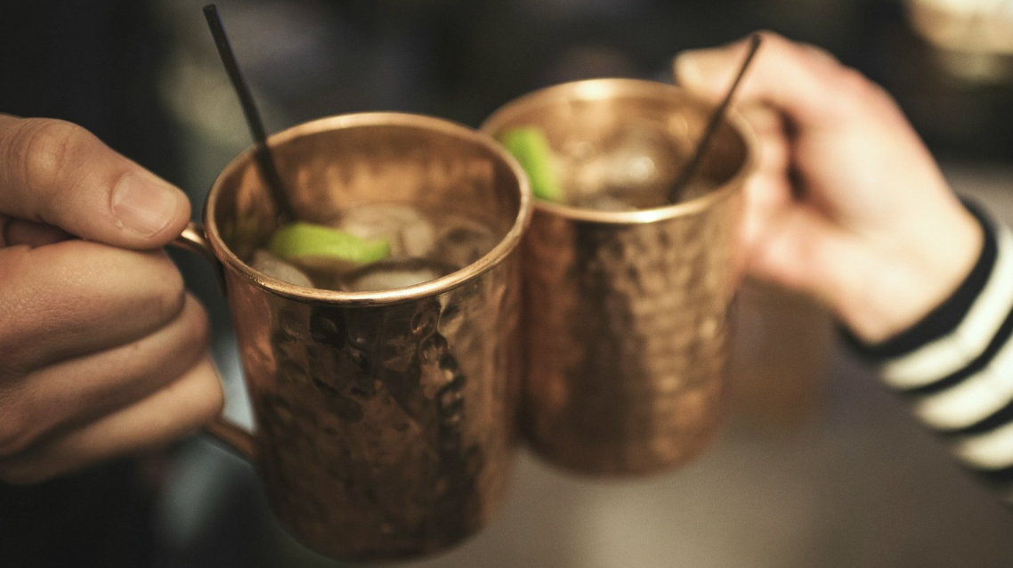 Two people are cheering with copper mugs filled with Bärenfang Mules with limes, ice, and black straws