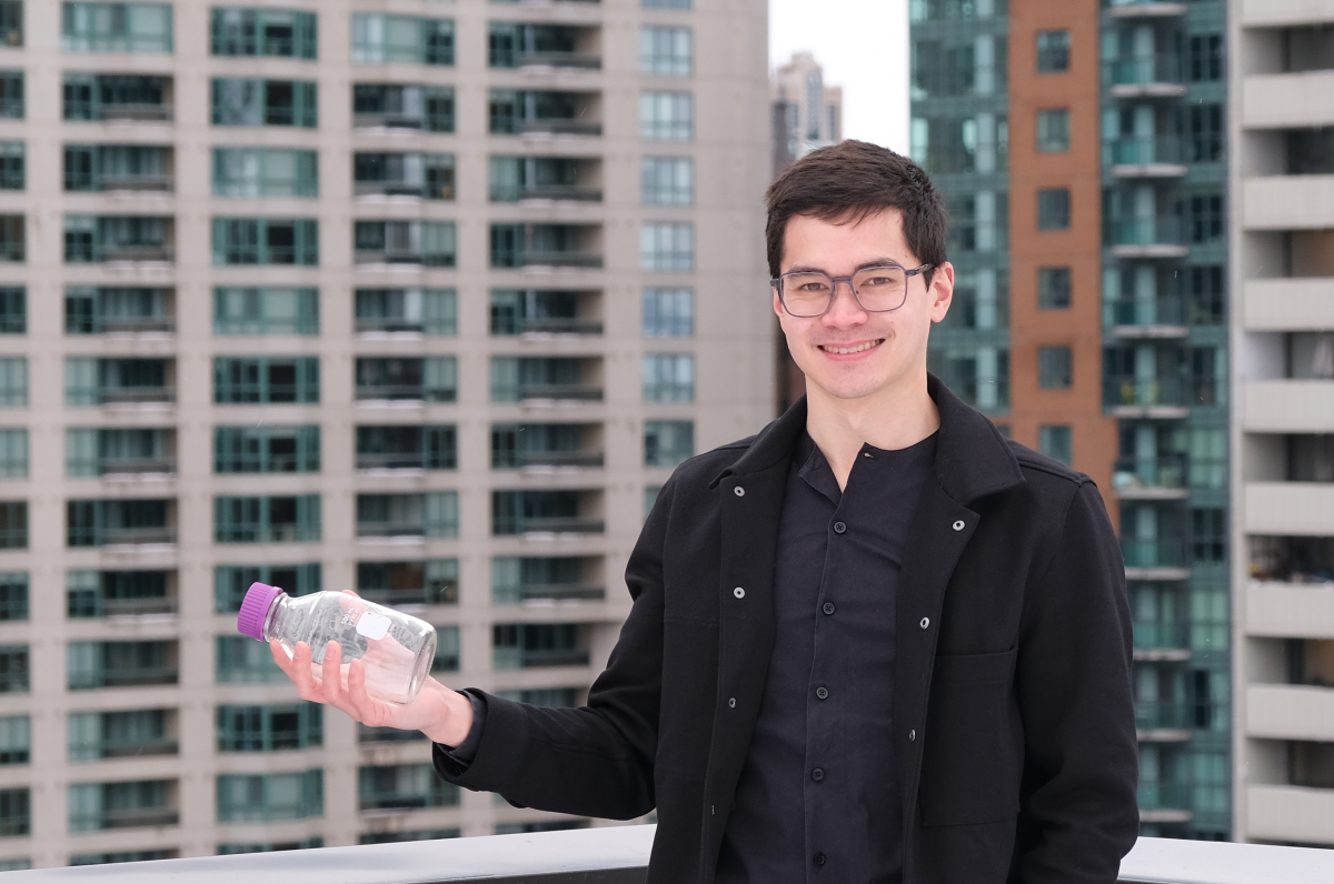 Kevin Chen wearing a black shirt, black jacket and grey glasses, holding a glass bottle standing on the roof of a building.