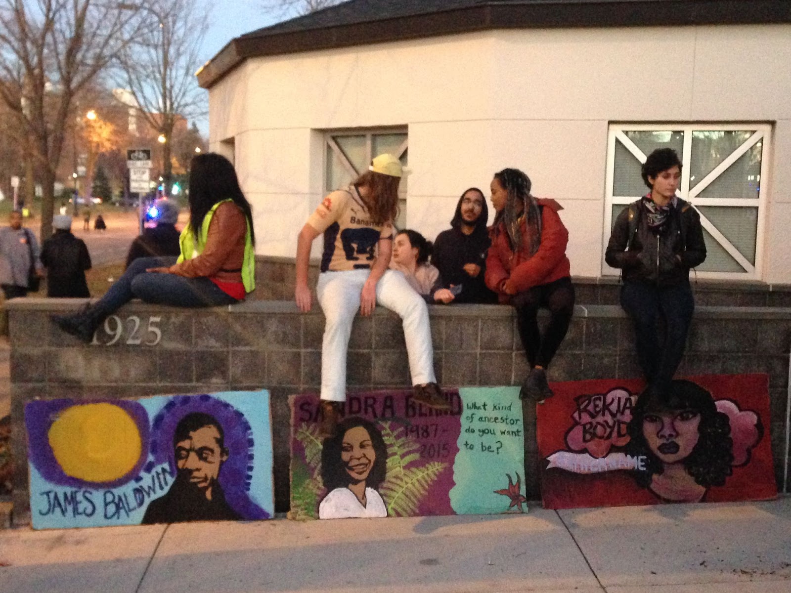 Image: Activist gathering outside the 4th Precinct during the Jamar Clark occupation, 2015. Several folks sit on a wall in front of the 4th Precinct building with works of art sitting right below them that depict folks like James Baldwin. Photo by the author.