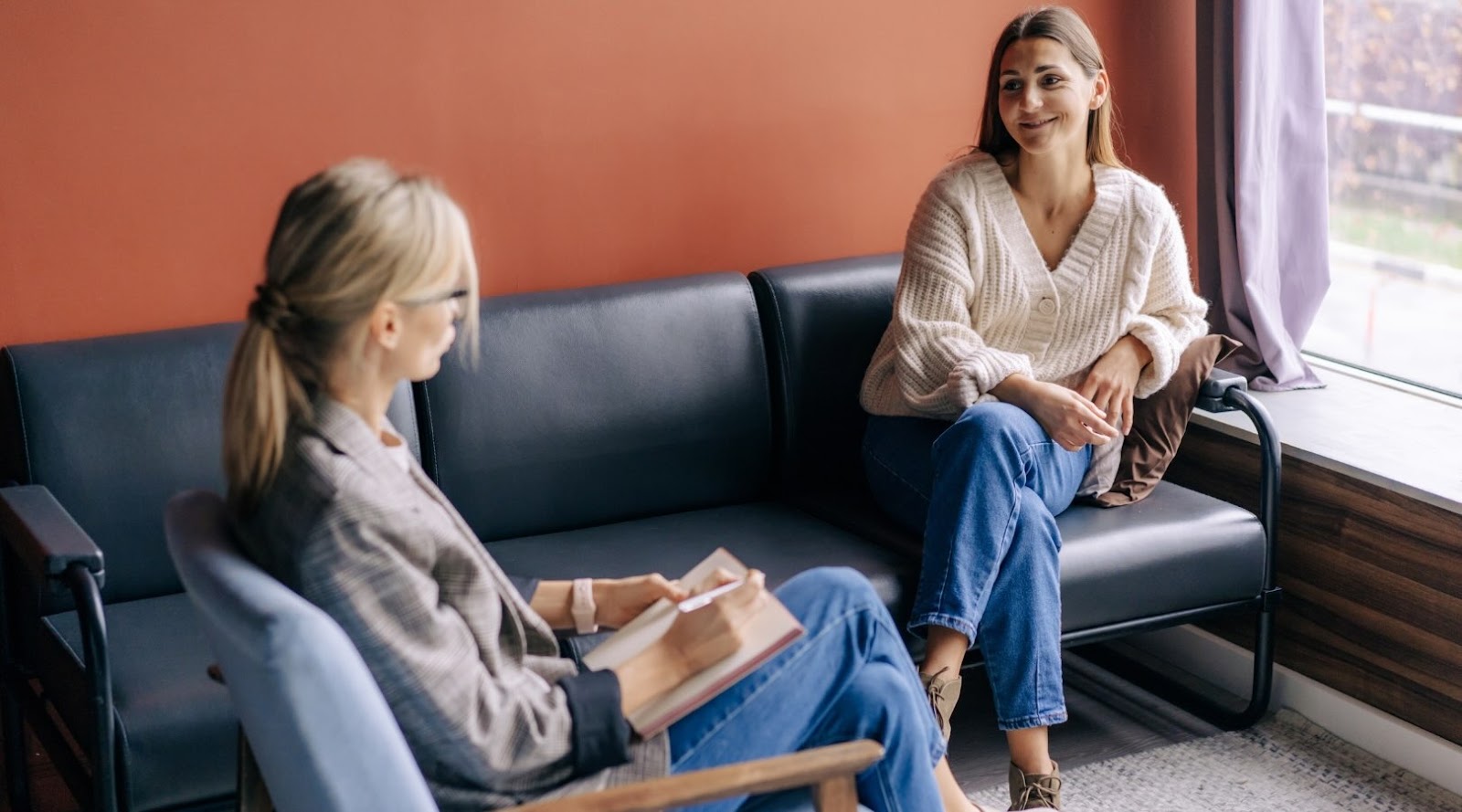 A smiling woman, dressed in a comfortable white sweater, is seated on a couch near a window, engaging in a conversation with a coach or professional therapist. The coach, wearing glasses and a plaid blazer, is sitting across from her, taking notes in a notebook. The atmosphere is warm and welcoming, suggesting a relaxed and supportive environment.