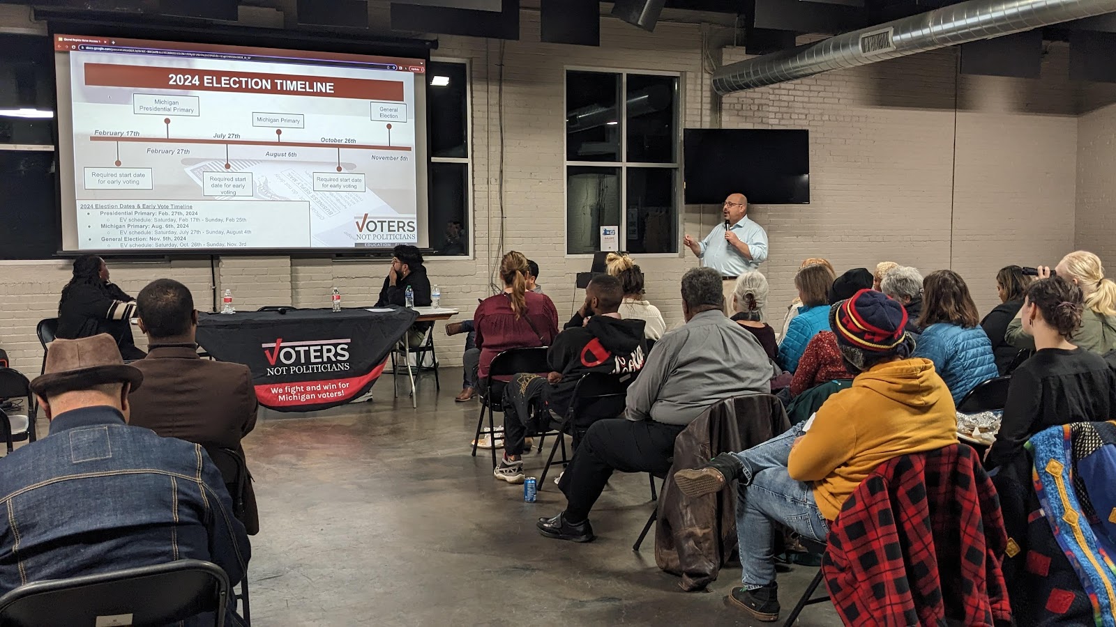 Community meeting with a presenter discussing the ''2024 Election Timeline'' on a projected screen, around twenty people are visible in the audience.