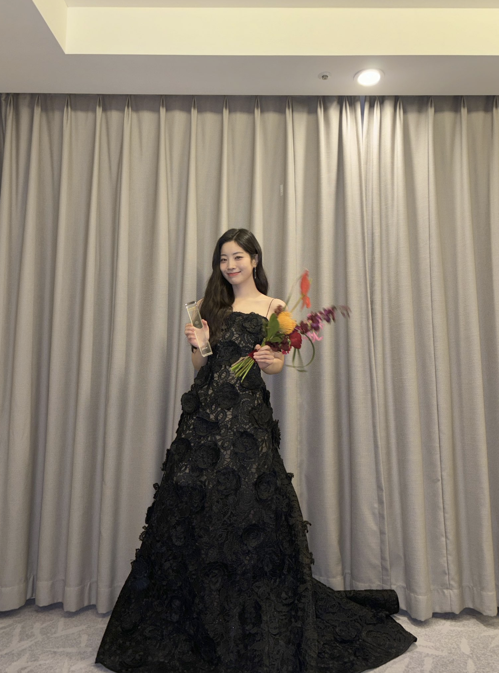 A picture of TWICE's Dahyun wearing a daring backless dress at the 2024 Marie Claire Korea Asia Star Awards during the Busan International Film Festival (BIFF) with her award in right hand and a flower on her left hand.