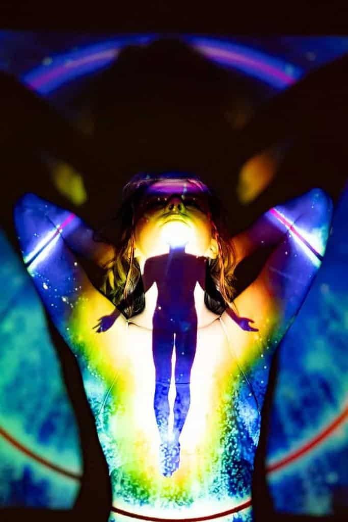 An individual is standing with a colorful, abstract projection on their body. The projection includes vibrant hues of blue, green, yellow, and purple, creating a visually striking effect. At the center of the projection, there is a silhouette of a person with arms outstretched, adding an intriguing element to the composition. The overall effect is artistic and visually captivating.