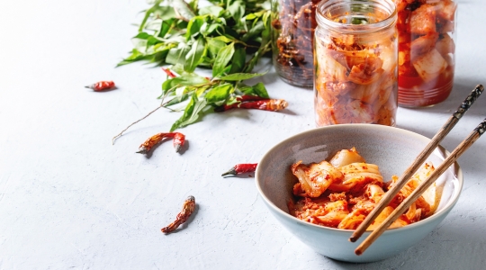 Un bol de kimchi avec des baguettes, entouré de piments séchés et de pots de kimchi en arrière-plan.