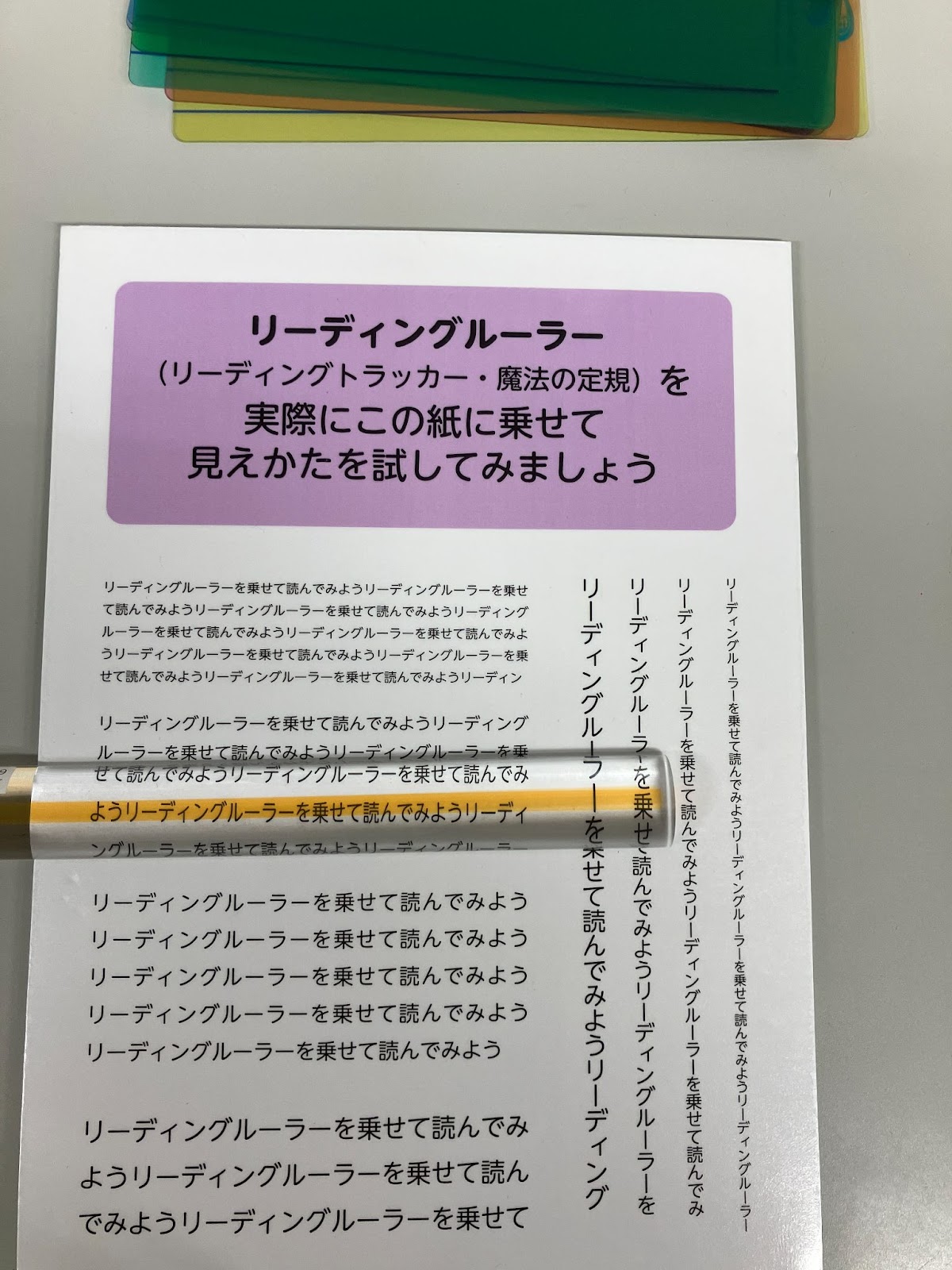 この画像は「リーディングルーラー」（リーディングトラッカー・魔法の定規）の説明と実際に試してみるための用紙です。カラフルなリーディングルーラー（透明な定規）が上に置かれており、文章を追いやすくするために使用されることが示されています。「実際にこの紙に乗せて見え方を試してみましょう」と書かれており、リーディングルーラーを実際に体験できるようになっています。