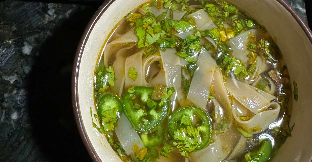 (Bowl of pho with wide rice noodles, sliced jalapeños, chopped cilantro, and a clear, golden broth served in a ceramic bowl on a dark countertop.)

