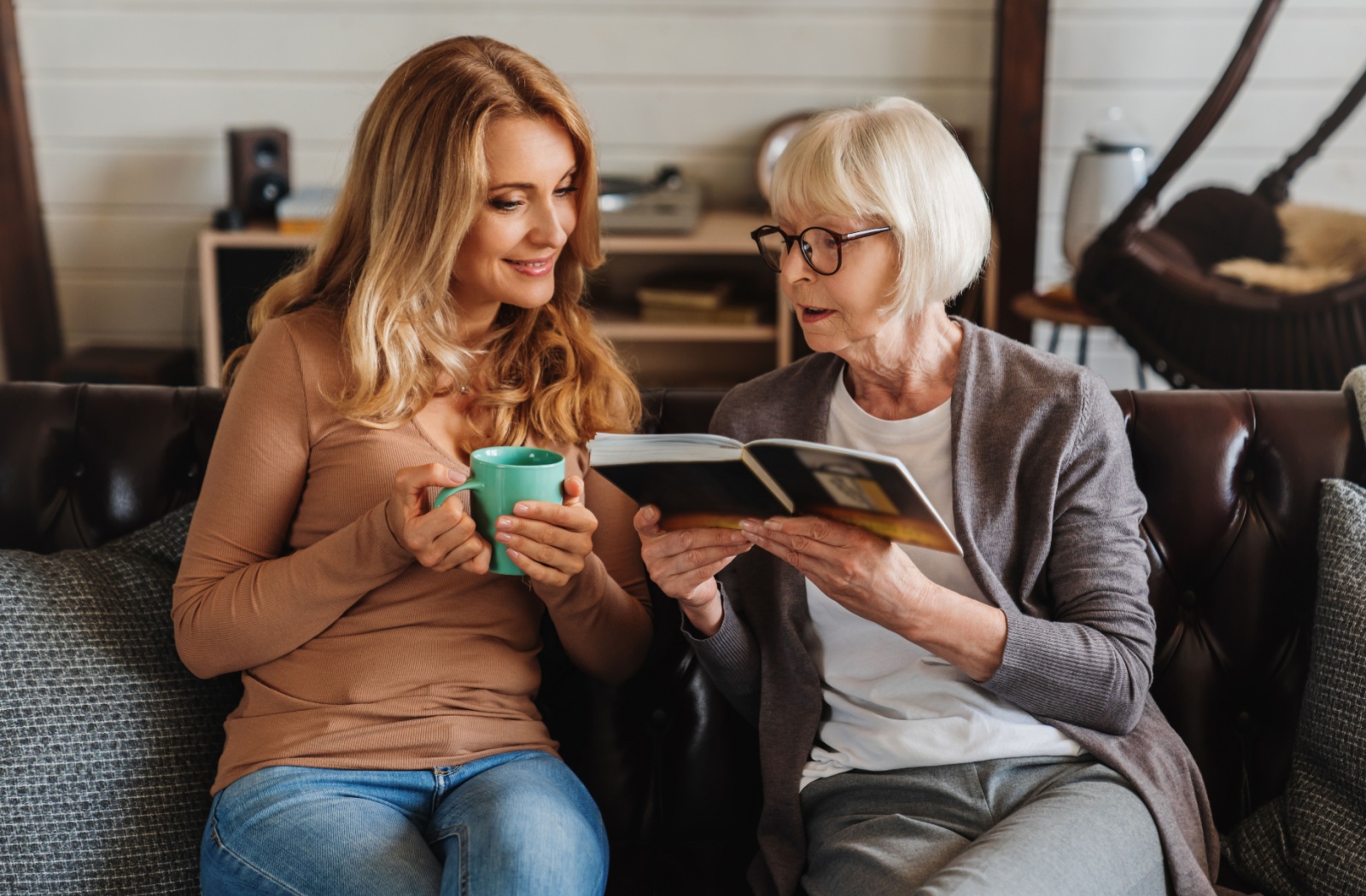 An elderly mother looking at a photo album with her young daughter, who is sipping a cup of coffee.