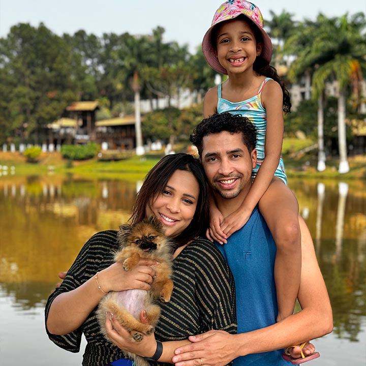 Família feliz ao ar livre com uma cachorrinha, crianças e adultos sorridentes, cenário de parque com lago e árvores ao fundo.