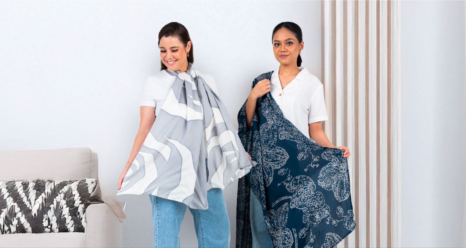 An image showing two women is holding and styling batik scarves from Batik Boutique in Grey Rose and Navy Orchid with the background of home.