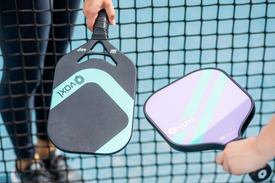 Close-up of two Voxl pickleball paddles held near the net, showcasing paddle designs and court detail.