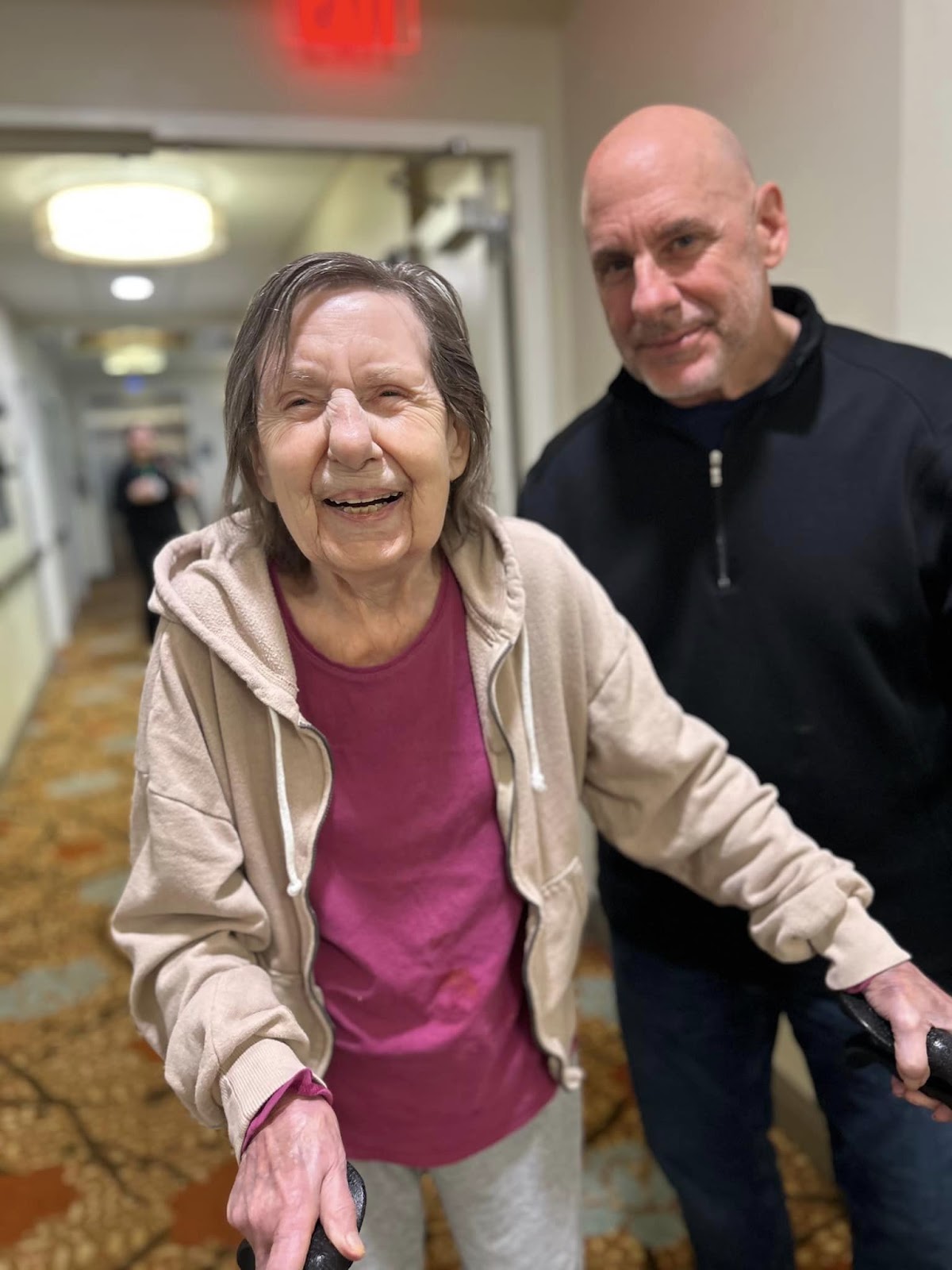 An elderly woman smiling while walking with a walker through the hallway of a memory care facility