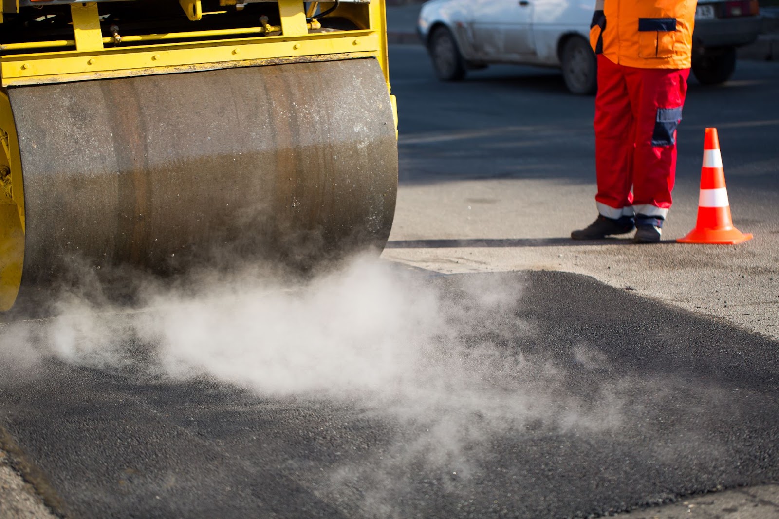 A road roller compacts fresh asphalt as a worker stands beside a safety cone.